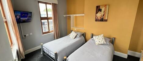 A room with two single beds, a TV mounted on the wall, a window with curtains, and a painting on the wall.