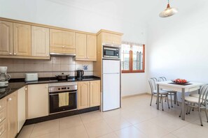 Una cocina con armarios de madera, estufa, horno, microondas y refrigerador. Hay una mesa de comedor con sillas y una ventana con cortinas.