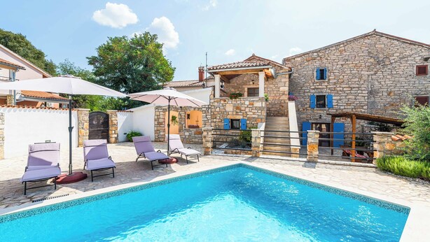 A swimming pool with lounge chairs and umbrellas, stone buildings, and a clear sky.