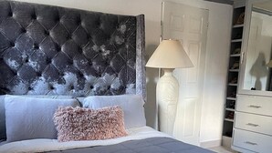 Een slaapkamer met een gestoffeerd hoofdeinde, een grijze spreidek, een zacht roze kussen, een witte lamp en een witte garderobe met meerdere lades.