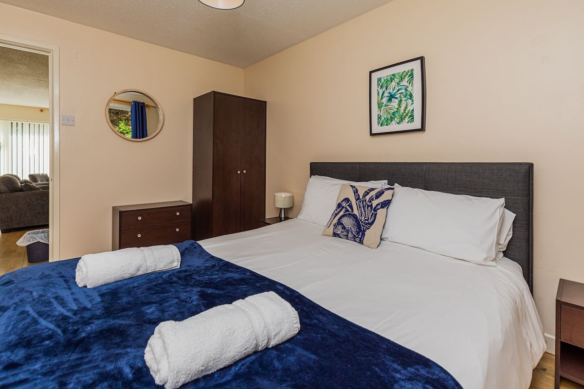 A neatly made bed with white and blue bedding, a dark wooden wardrobe, a round mirror, and a framed picture on the wall.