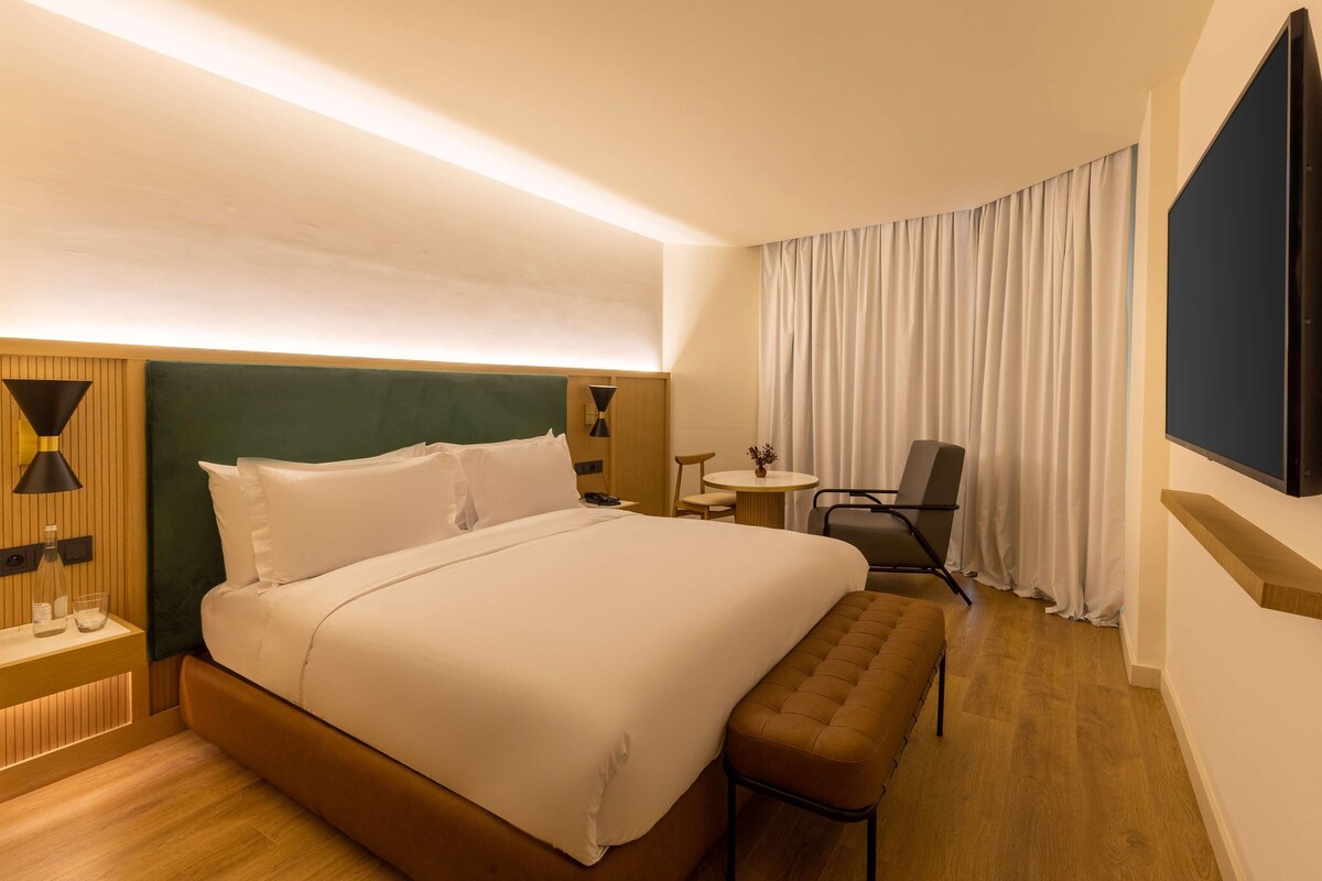Ein modernes Hotelzimmer mit einem großen Bett, einem kleinen runden Tisch, einem Stuhl und einem an der Wand befestigten Fernseher.