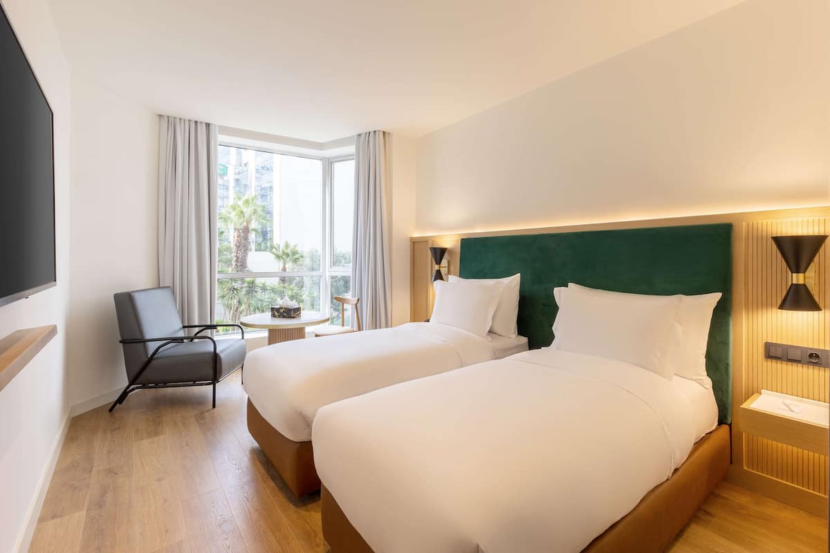 Ein Hotelzimmer mit zwei Betten, einem Sessel, einem kleinen Tisch und einem Fenster mit Vorhängen.