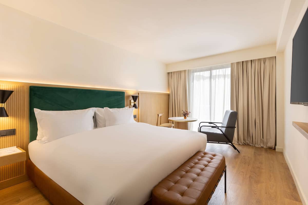 Ein Hotelzimmer mit einem großen Bett, einem Nachttisch, einem Sessel, einem kleinen runden Tisch und einem an der Wand befestigten Fernseher.