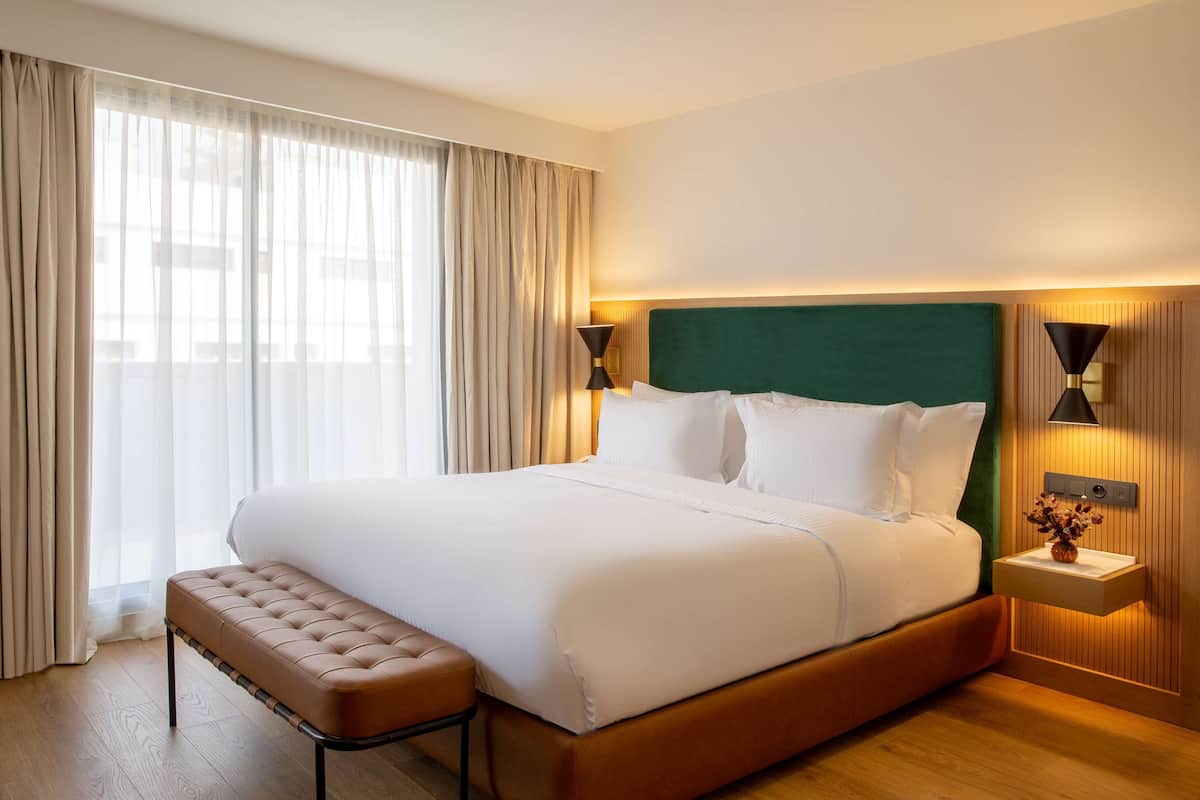 Ein Hotelzimmer mit einem großen Bett, einer Bank, einem Nachttisch mit Lampe und Blick ins Freie.