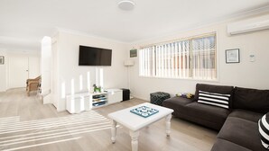 A modern living room with a flat-screen TV, a sofa, a coffee table, and a window with blinds. - Bombora Walking Distance to Fingal Beach- Farm Rd, Fingal Bay (Fingal Bay)