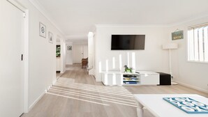 A modern living room with a flat-screen TV mounted on the wall, a white entertainment unit, and a window with blinds. - Bombora Walking Distance to Fingal Beach- Farm Rd, Fingal Bay (Fingal Bay)