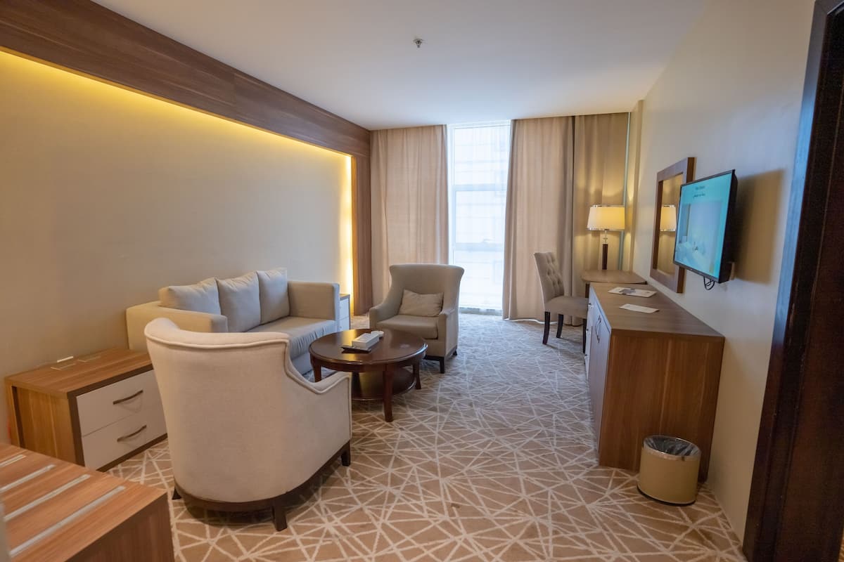 A hotel room with a sofa, armchairs, a small table, a TV, and a desk with a lamp.