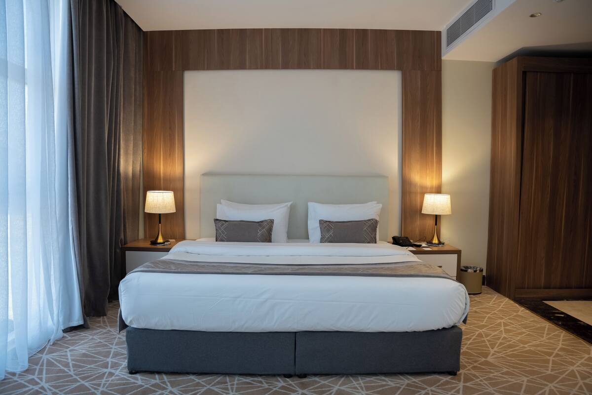 A hotel room with a large bed, two bedside tables, a headboard, and a window with curtains.