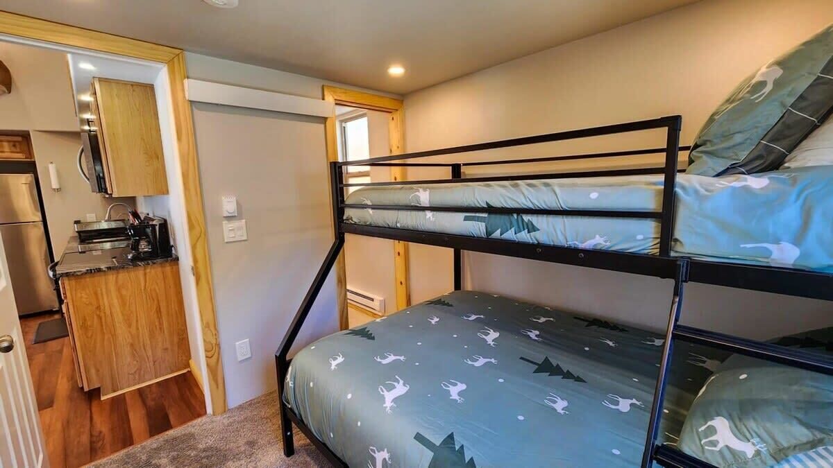 A bunk bed with a patterned bedspread, a kitchenette with wooden cabinets, and a door leading to another room.