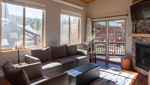 A cozy living room with a stone fireplace, a sofa, and large windows offering a view of a wooden deck and trees. - 2BR Riverside Cabin - Near Trails w/Hot Tub Access (Breckenridge)