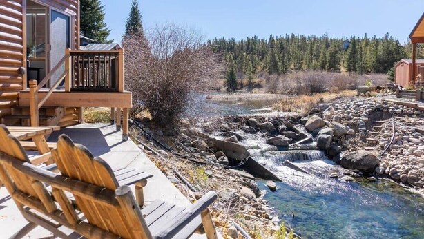 A wooden cabin with a deck overlooking a clear stream and forest. - 2BR Riverside Cabin - Near Trails w/Hot Tub Access (Breckenridge)