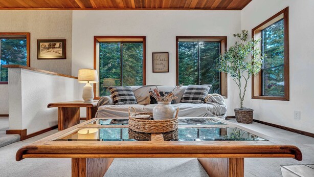 A cozy living room with a wooden ceiling, large windows, a sofa with plaid cushions, a glass coffee table, and a potted plant.