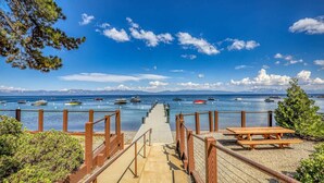 A wooden boardwalk leading to a dock with boats, a picnic table, and mountains in the background.