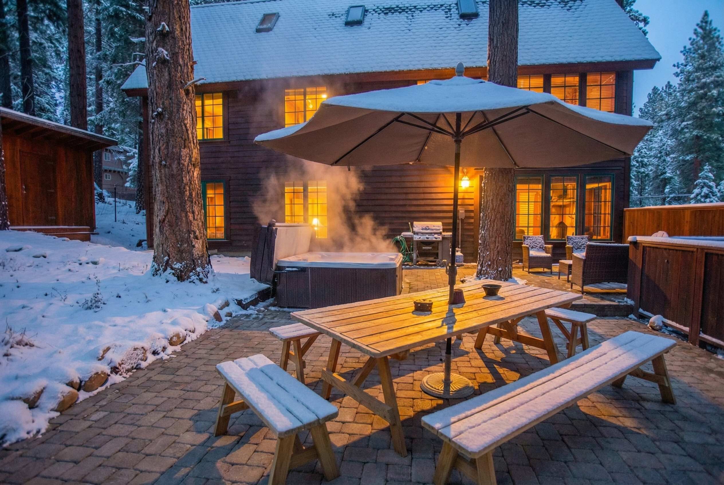 Une cabane en bois dotée d’un patio comprenant une table et des bancs, un jacuzzi et un grand parasol.