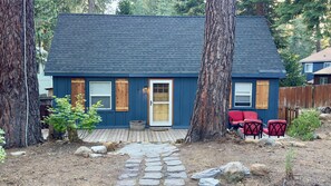 A house with a dark blue exterior, a front door, two windows, a wooden deck, and a small garden area with rocks and a potted plant.