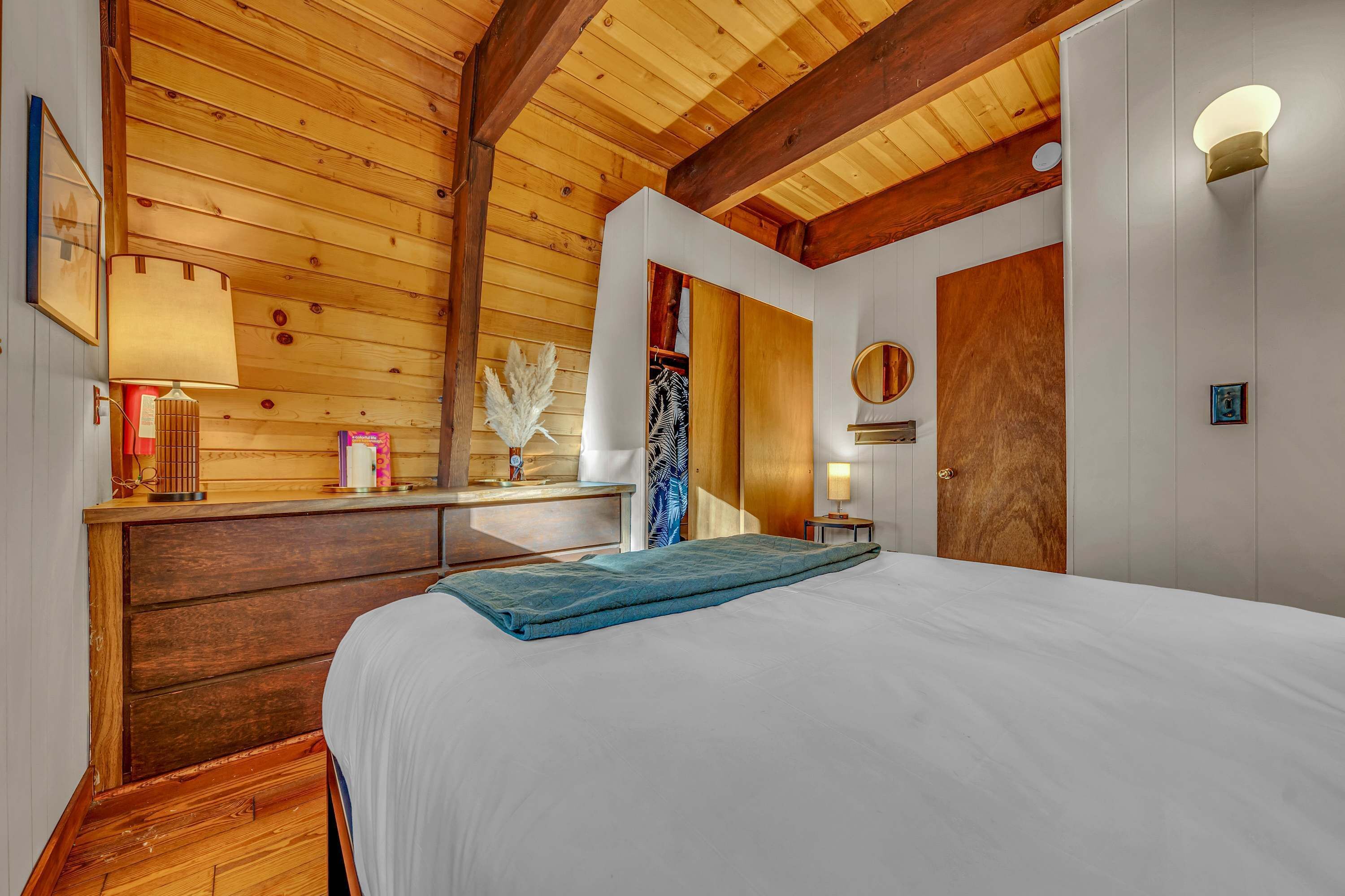 Ein Schlafzimmer mit Holzboden, einem Bett mit blauer Decke, einer hölzernen Kommode, einem Nachttisch mit Lampe und einem Kleiderschrank.