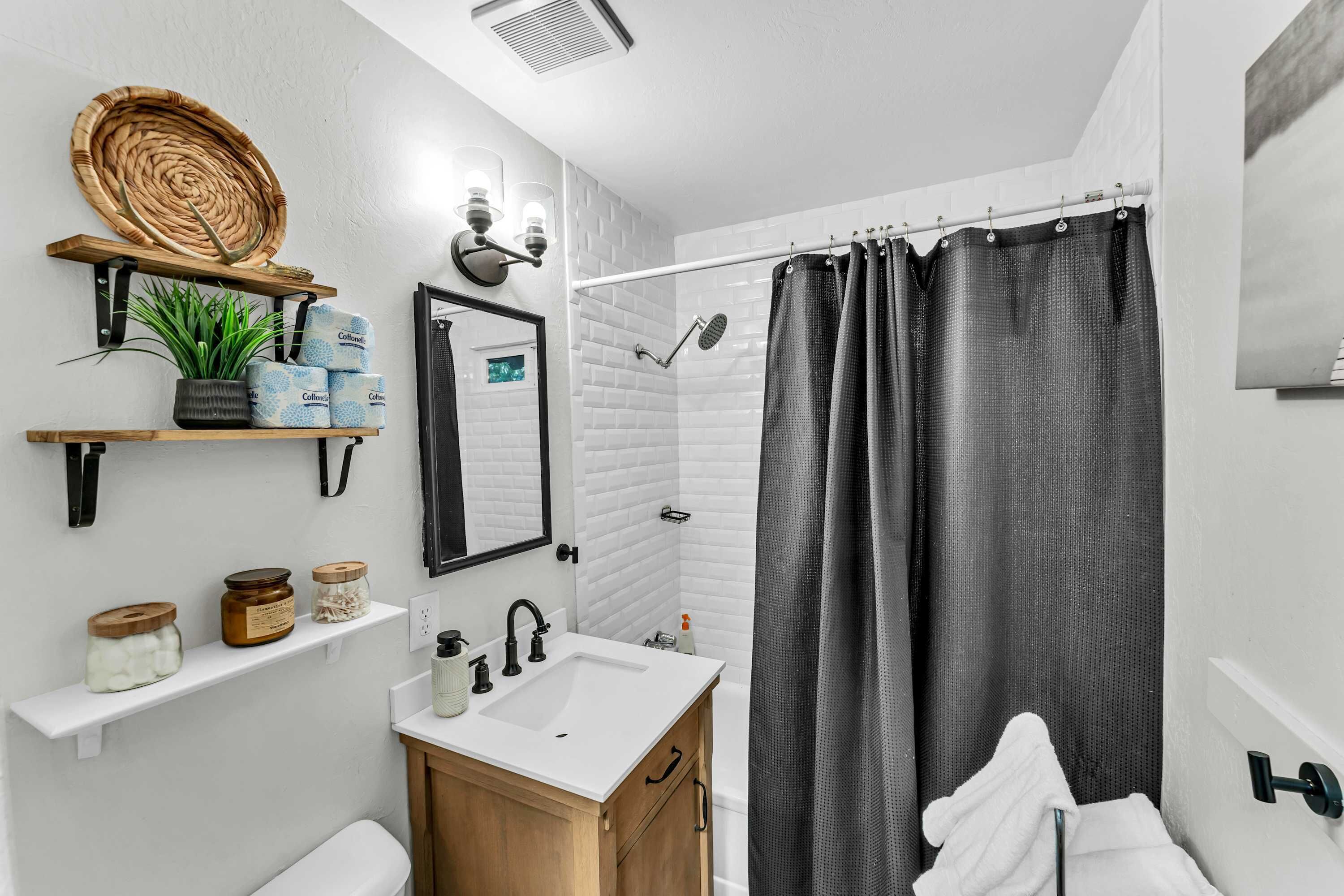 A modern bathroom with a white sink, wooden vanity, and a shower with a black curtain. There are shelves with plants and toiletries.