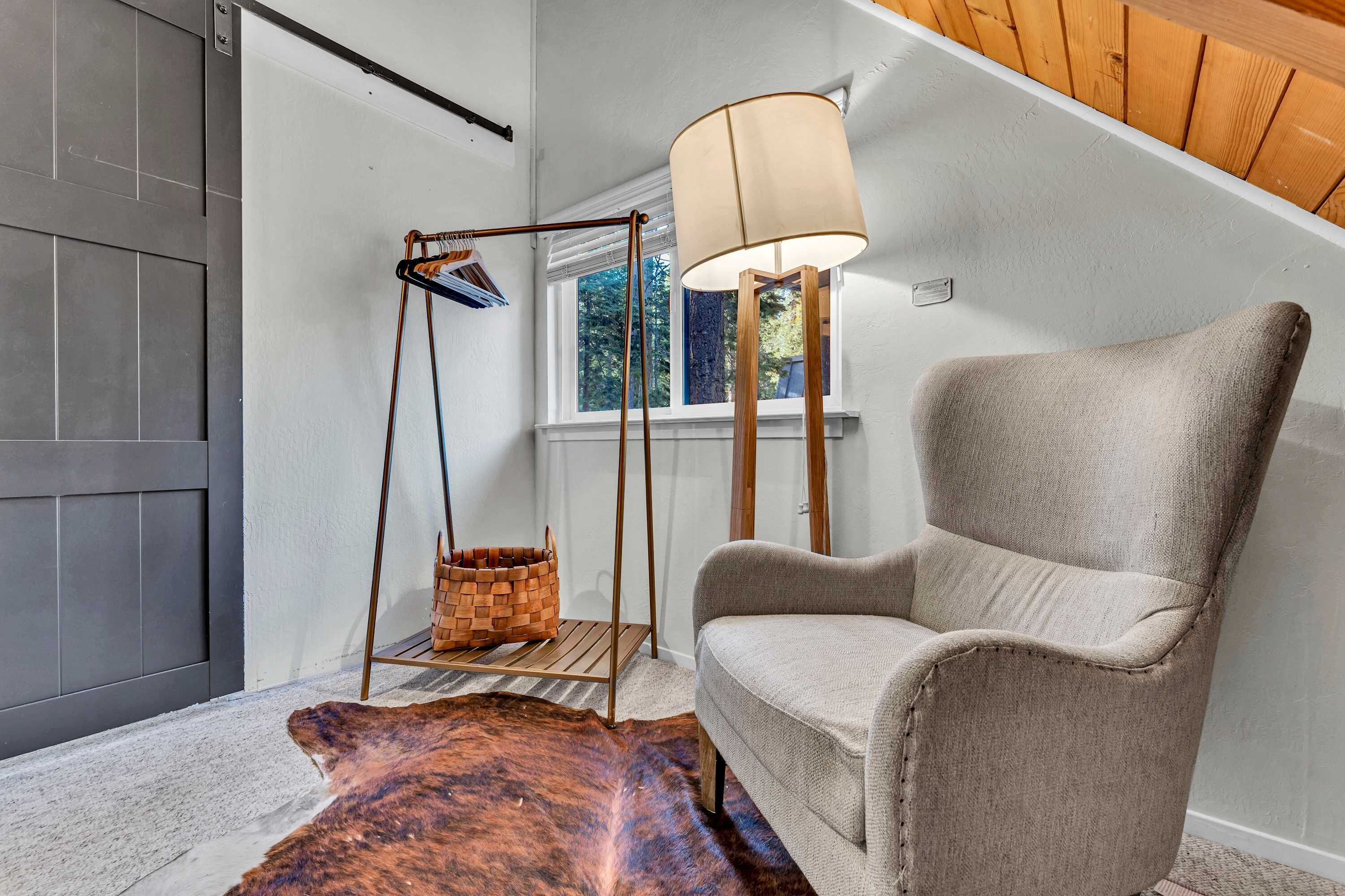 Een kamer met een grijze fauteuil, een houten vloerlamp, een hangmand en een groot raam met uitzicht op bomen.