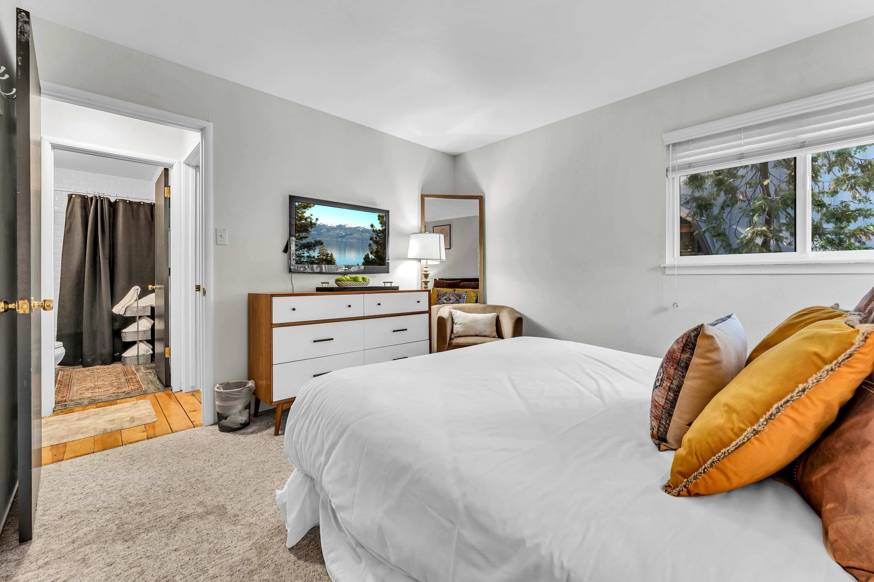 A bedroom with a bed, a dresser, a mirror, a window, and a door leading to a bathroom.