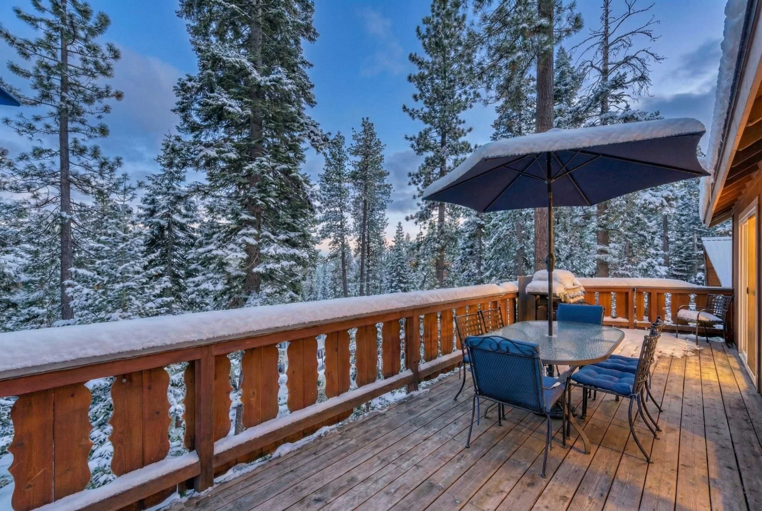 A wooden deck with a table and chairs, surrounded by snow-covered trees.