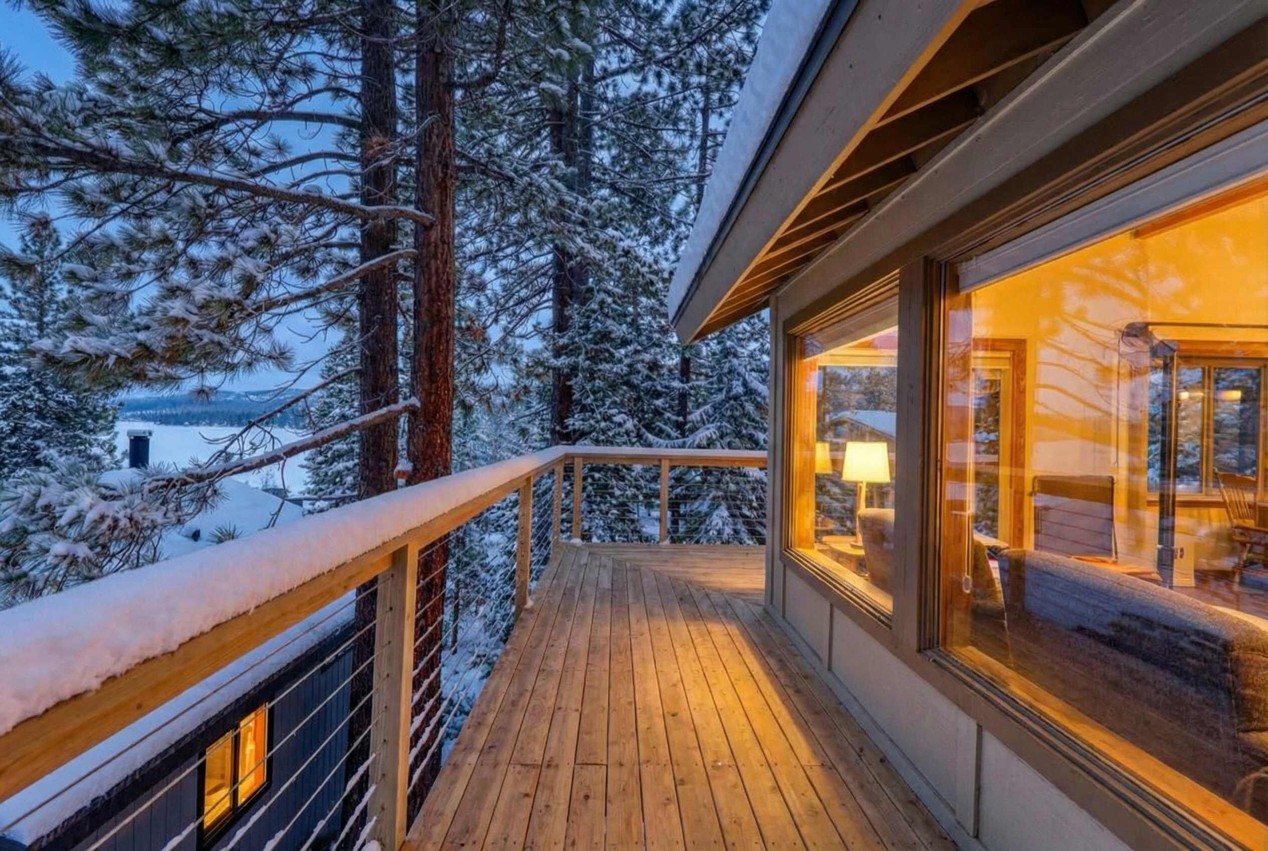Une terrasse en bois avec une balustrade, un intérieur chaleureux éclairé par une lampe, et un paysage enneigé en arrière-plan.