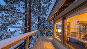 A wooden deck with a railing, a cozy interior with a lamp, and a snowy forest backdrop. - Tahoe Vista Retreat with Lake View- Walk to beach! (Tahoe Vista)
