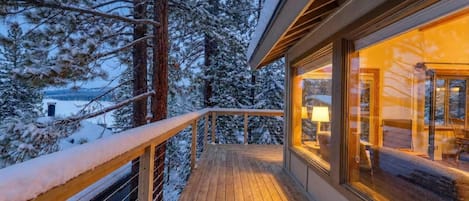 En træterrasse med gelænder, et hyggeligt interiør med en lampe, og en sneklædt skov i baggrunden.