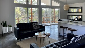 En rummelig opholdsstue med en sort sofa, et sofabord og et spiseområde med barstole. Store vinduer giver udsigt til det fri.