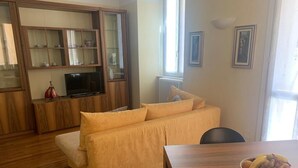 Una sala de estar con un sofá, un televisor sobre un mueble bajo, una mesa de comedor con fruteros y una ventana con cortinas.