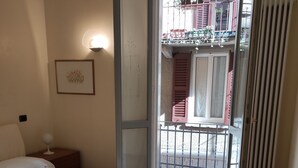 Habitación de hotel con cama, mesita de noche con lámpara, banco y vista al balcón exterior.
