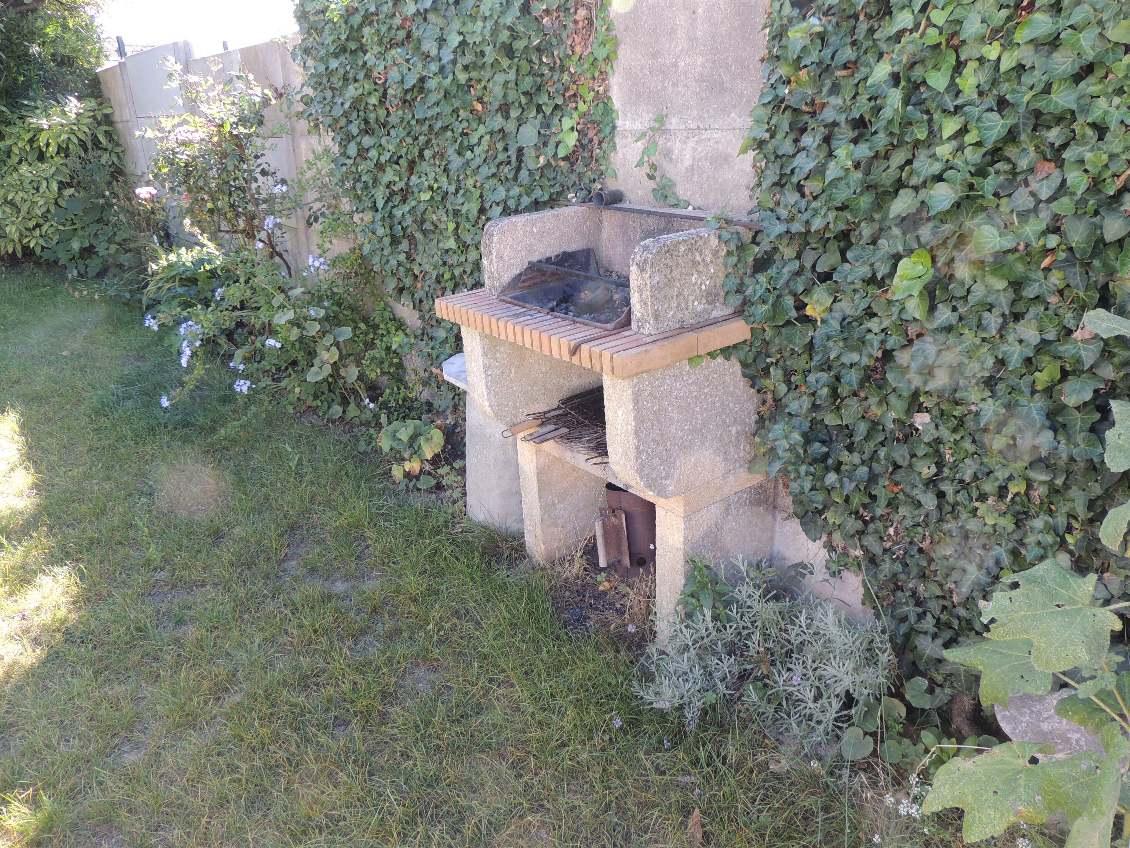 Un barbecue extérieur en béton, surmonté d’une surface en briques, entouré de verdure et bordé d’un mur de pierres.
