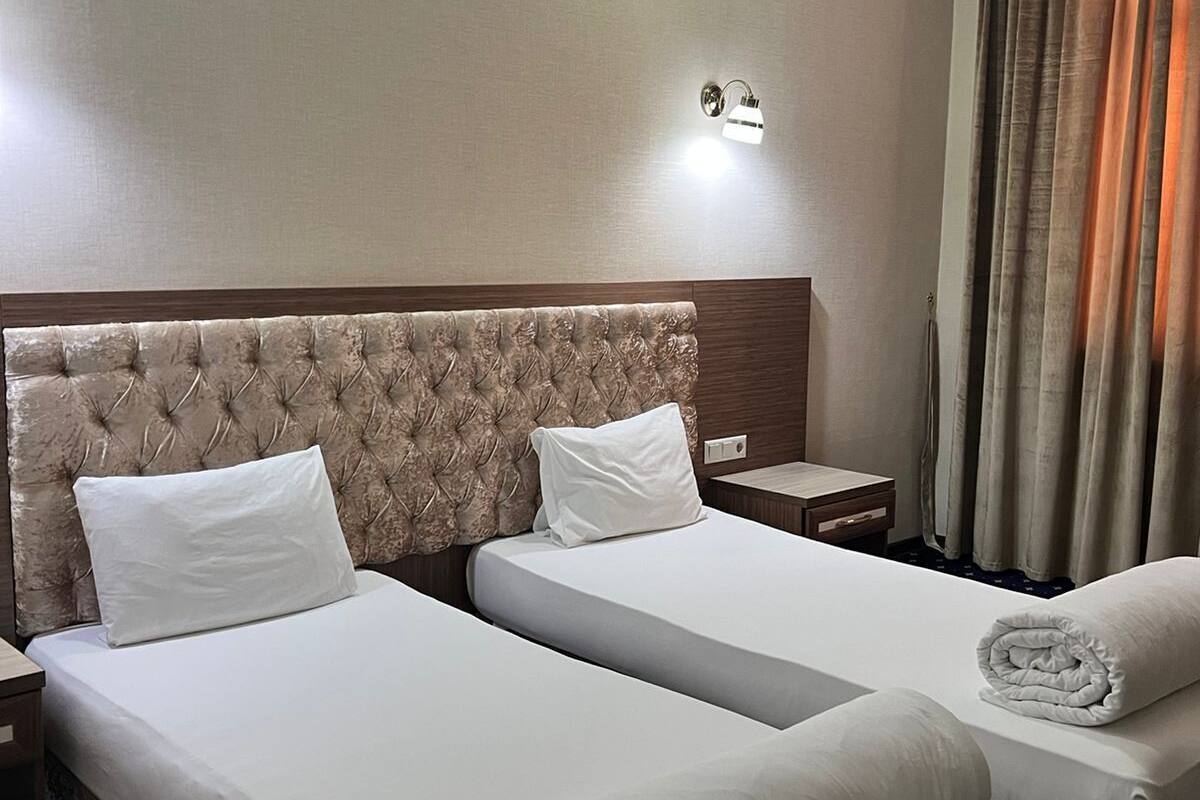 A hotel room with two beds, a nightstand, a wall-mounted lamp, and a window with curtains.