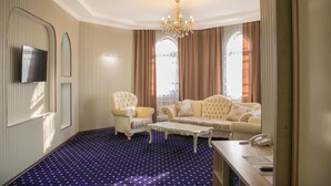 A hotel room with a large window, a chandelier, a sofa, a chair, a television, and a patterned carpet. - Hotel Khanaka (Turkestan)