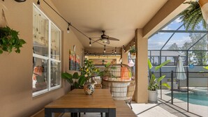 Un patio couvert avec une table en bois, des plantes en pot, un ventilateur de plafond et une vue sur une piscine.