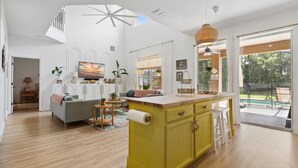 Une cuisine lumineuse avec un îlot central jaune, un espace de vie comprenant un canapé et un téléviseur, et une grande fenêtre donnant sur l’extérieur.