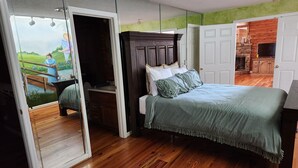 A bedroom with a large bed, a dresser, and a mirror. There is a view of a kitchen and a dining area through an open door.