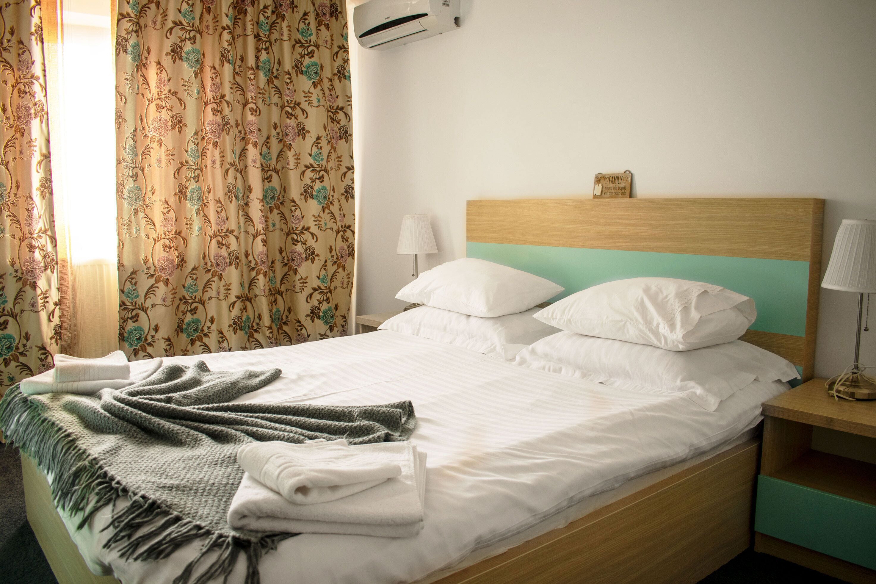 Una cama bien hecha con sábanas blancas y una manta verde, cabecera de madera, lámpara de noche y aire acondicionado instalado en la pared.