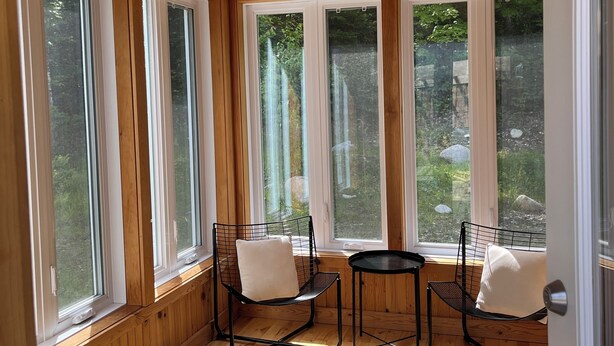 Una habitación con paredes y techo de madera, ventanas amplias, dos sillas de malla metálica negra con cojines blancos, una pequeña mesa redonda y una lámpara en el techo.