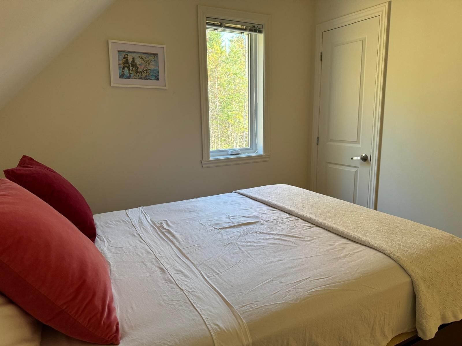 A bedroom with a bed, a window, a door, and a framed picture on the wall.