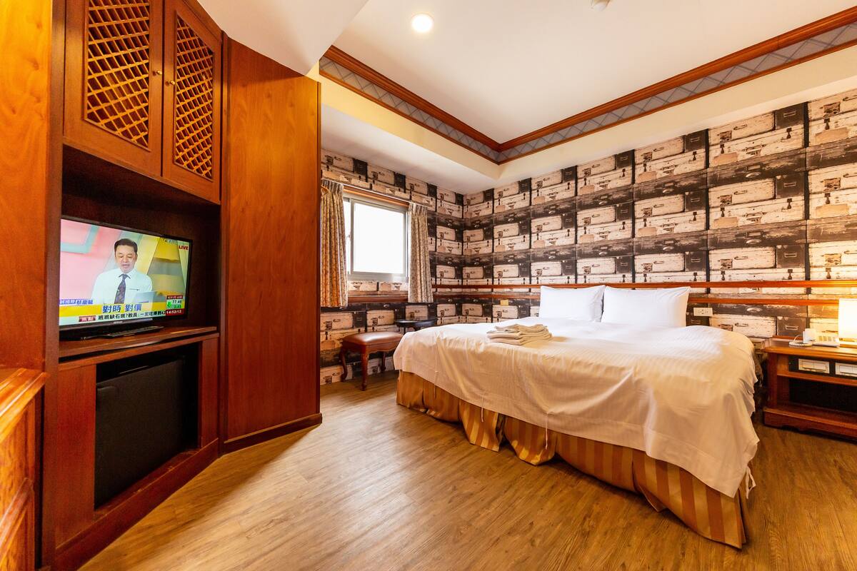 A hotel room with a wooden floor, a bed with white bedding, a television on a wooden stand, and a wall with a patterned design.