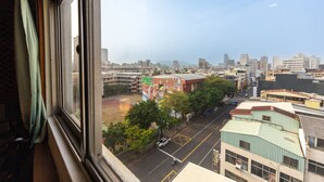 Une vue sur un paysage urbain, prise depuis une fenêtre, qui présente un mélange de bâtiments modernes et plus anciens, un ciel dégagé et une rue où l’on voit des véhicules et des arbres.