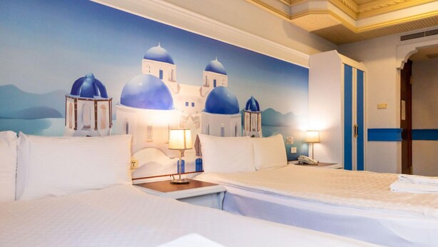 Une chambre d’hôtel avec une fresque représentant une île grecque, un lit, des tables de chevet et une petite table de nuit avec une lampe.