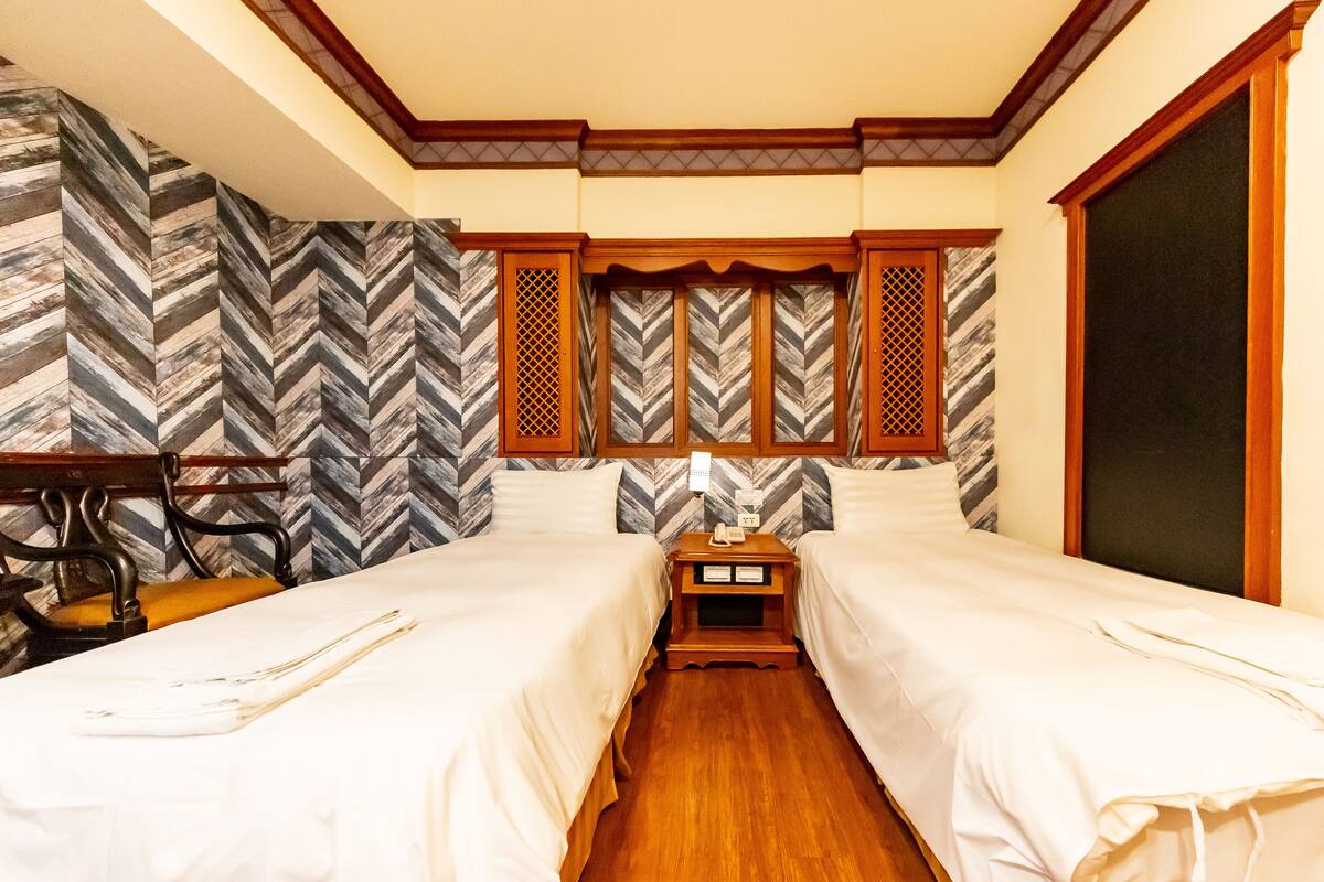 A hotel room with two beds, a desk, and a chair. There is a wooden floor and a large window.