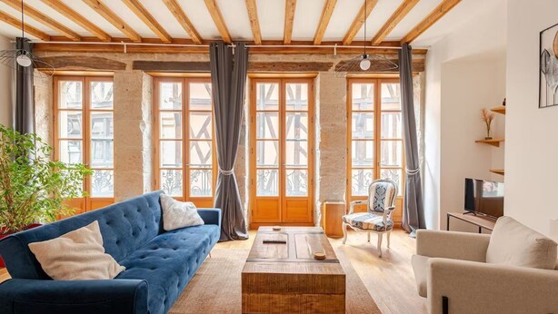 A cozy living room with a blue sofa, a wooden coffee table, and a white armchair. Large windows with curtains allow natural light to fill the space.