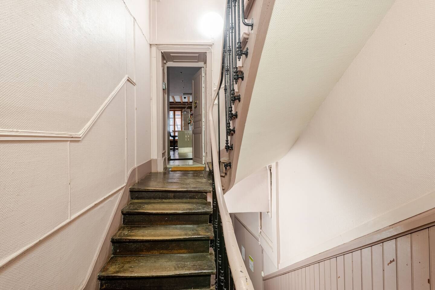 Uma escadaria estreita com corrimão de um lado e parede do outro, levando a uma porta no topo.