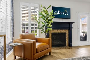 A living room with a brown leather sofa, a small side table, a large plant, a fireplace, and a television mounted on the wall.