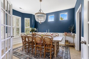 A dining room with a white table, wooden chairs, a chandelier, and a patterned rug.