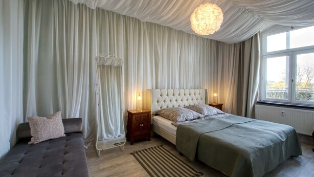 A bedroom with a tufted headboard, a bed with green bedding, a grey sofa, a white canopy bed, and a large window with a view. - Vacation apartment Herrenzimmer with fireplace and terrace (Annaberg-Buchholz)
