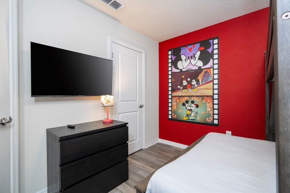 Ein Schlafzimmer mit Bett, Schminktisch, einem an der Wand montierten Fernseher und einer roten Akzentwand mit einem Mickey Mouse Wandbild.